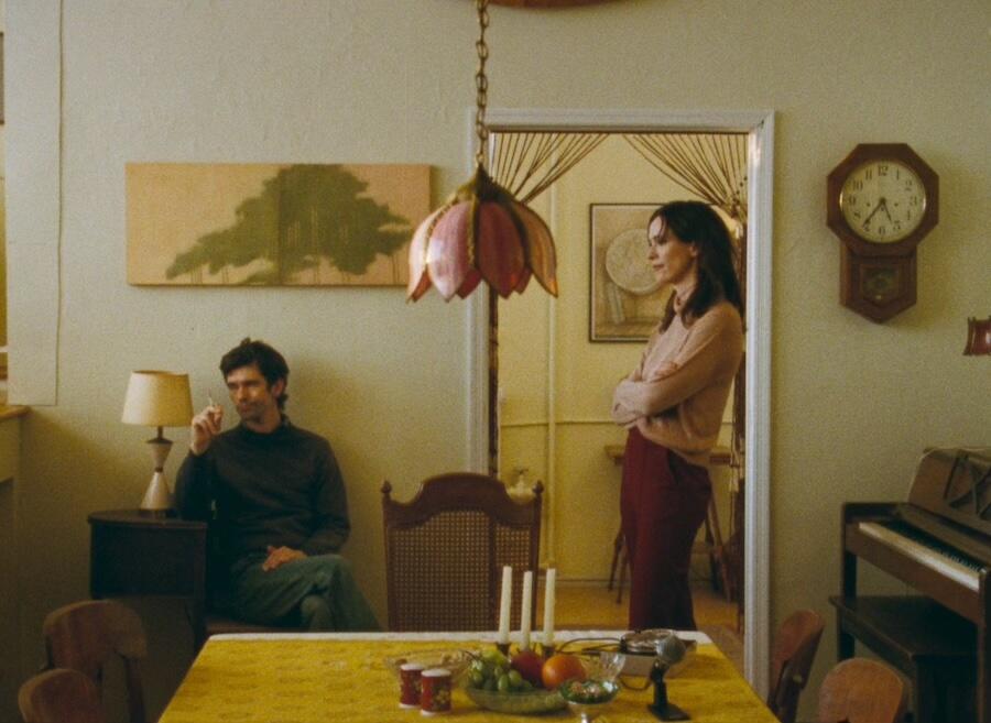 Ben Whishaw as Peter Hujar sits in a chair holding a cigarette and Rebecca Hall as Linda Rosenkrantz stands in profile looking into distance