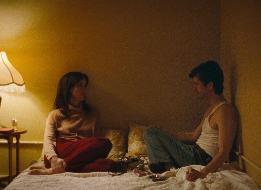 Ben Whishaw as Peter Hujar and Rebecca Hall as Linda Rosenkrantz sit on a bed, legs folded, in a dimly let room