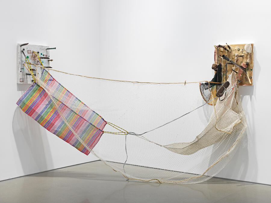 An installation view, uh, what looks to be a concrete block with metal and colorful components sticking out, which holds up a hammock that is strung across another corner against another... contraption