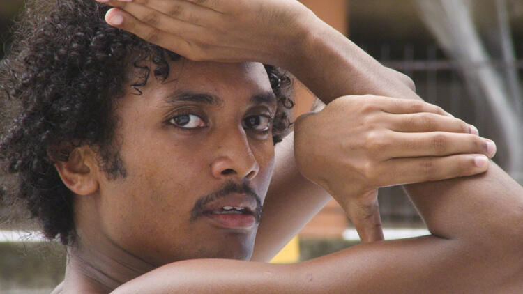 An image of a man with curly hair in the midst of a dance step: he turns toward his right and looks directly at the vier, his arm crooked at temple height, his other hand linked through it