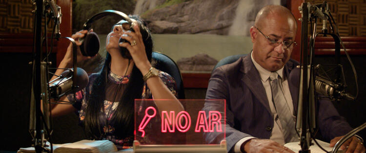 Two figures sit in what appears to be a recording studio; the words 'NO AIR' are in red bold letters upon a transparent block at the foreground. The female-appearing person on the left is midway between putting her headphones on or taking them off; the male-appearing person on the right wears a suit and appears to be writing something down