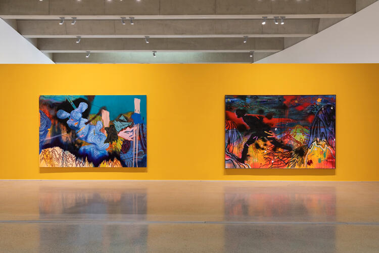 An image of two large-scale almost abstract paintings, the left mostly blue, the right a patchwork of black, orange, and blue, with a discernible black silhouette, against a yellow wall