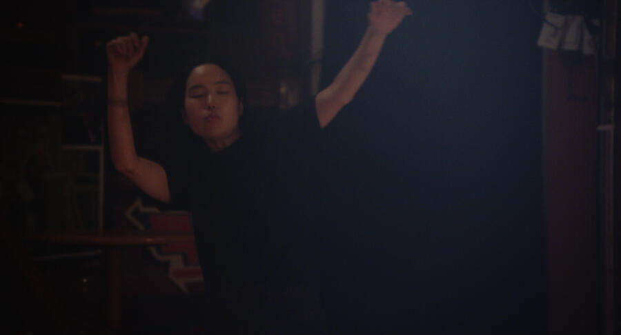 An Asian woman dances at a bar in a black T-shirt, eyes closed, arms overhead