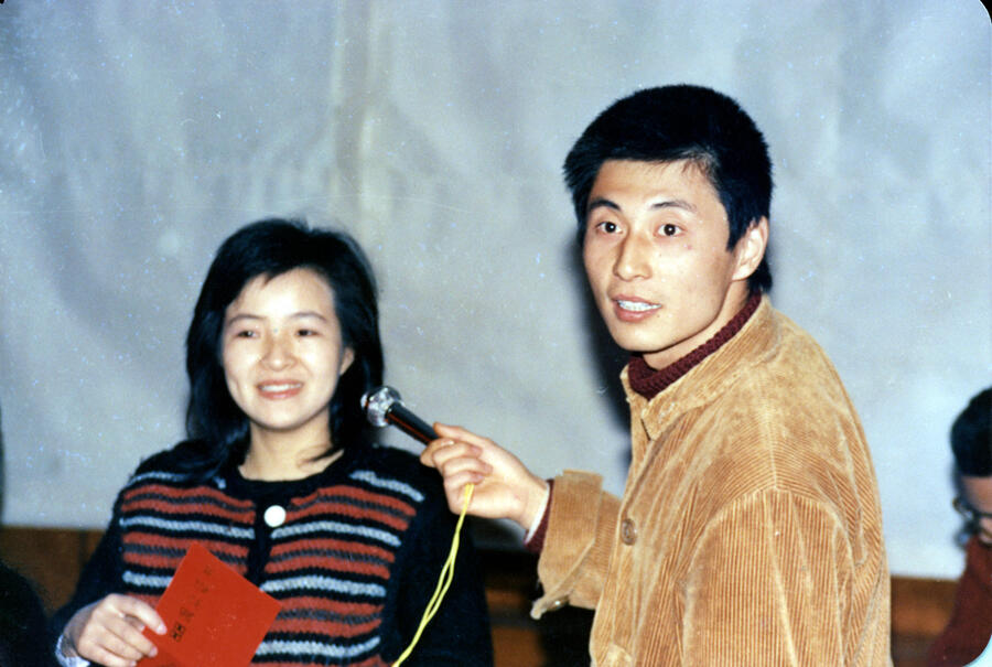 A documentary image of a man and woman: he, to the right, gazes somewhere behind to the left of the viewer, holding a microphone to his neighbor; she, left, smiles out