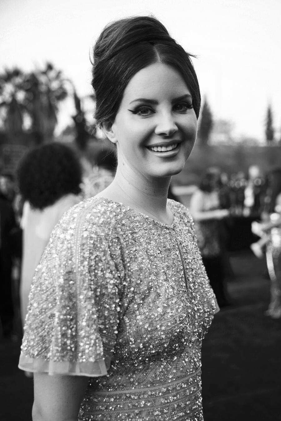 A black and white photograph of Lana Del Rey, her hair in an updo, wearing a sparkly dress, smiling