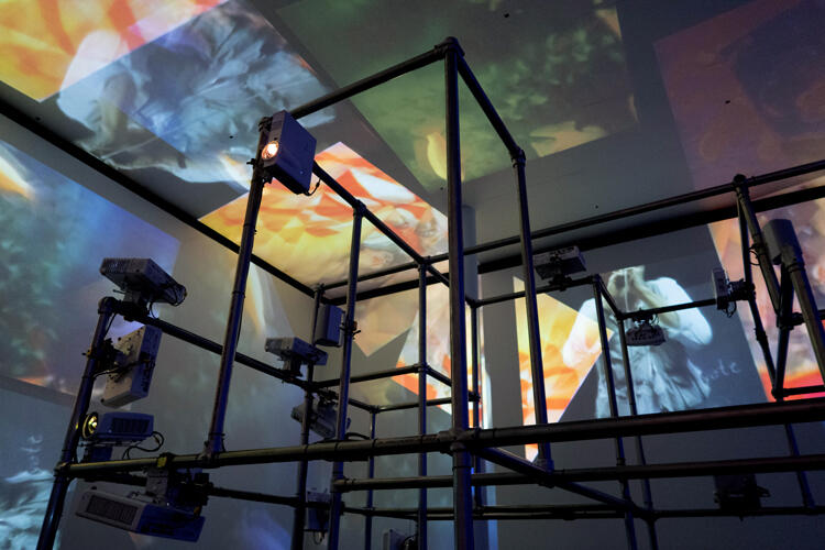 Image of a scaffold reaching up to the ceiling as miscellaneous projected image crisscross the walls, ceiling, and structure