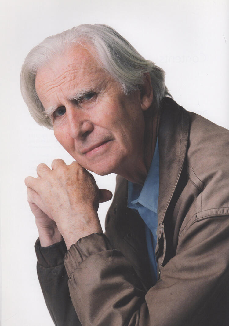 A photograph of Brian O'Doherty in old age: he has white hair and blue eyes, and peers over his left shoulder at us, chin gently resting on lightly clasped hands. He wears a light brown jacket and a blue button-up shirt with collar. 