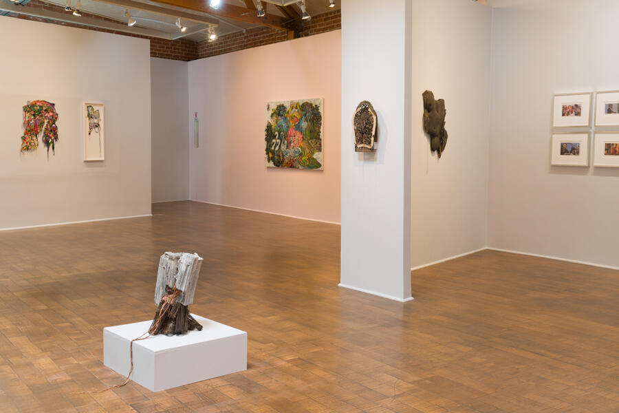 Installation view of a room with a wooden floor with a number of handicraft-esque objects: at the forefront, a brown and white object that trails from pedestal to floor